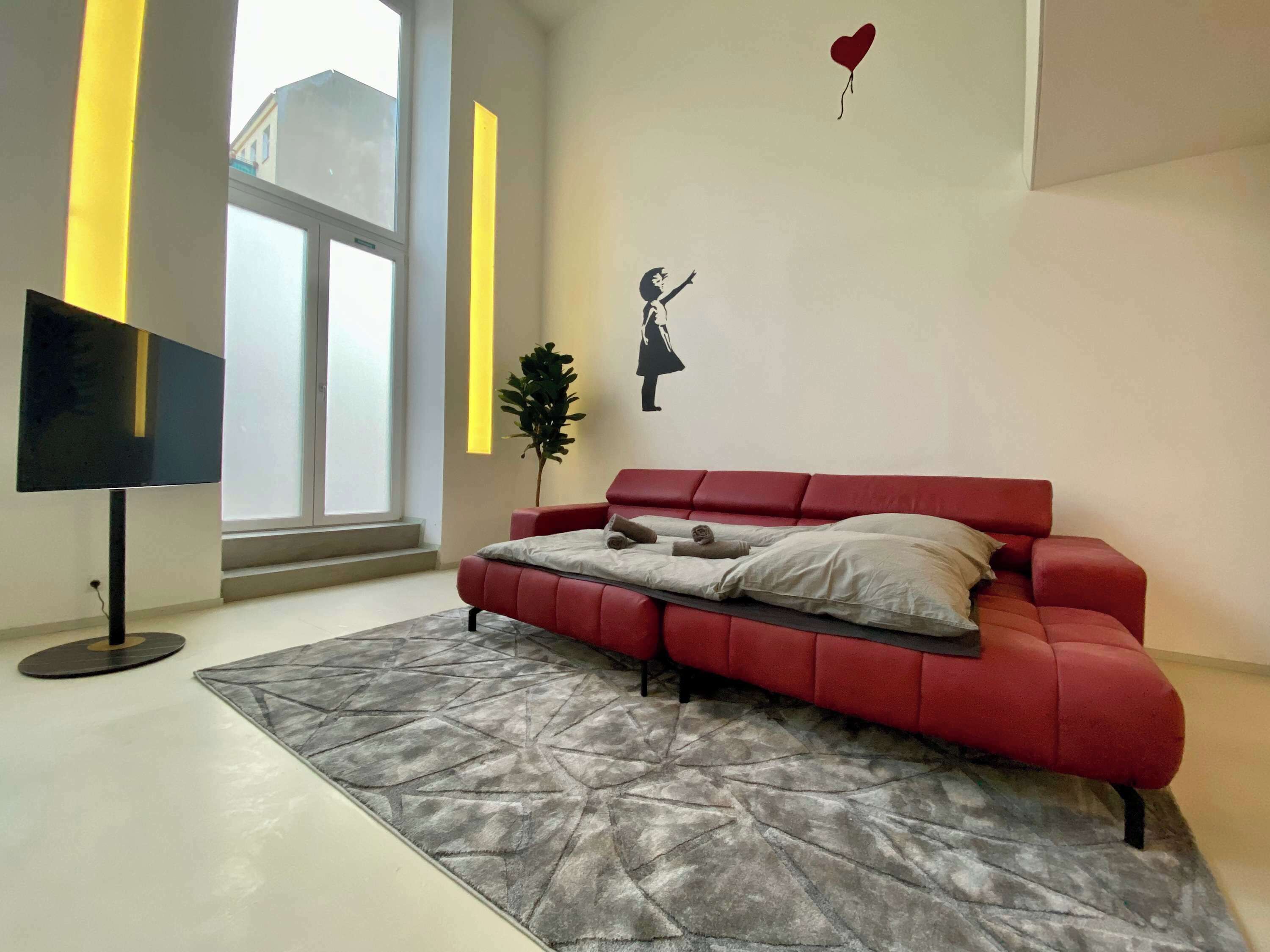 A modern living room with a red sofa, a grey patterned rug, a flat-screen TV, and a wall with a heart-shaped balloon and a silhouette of a person.