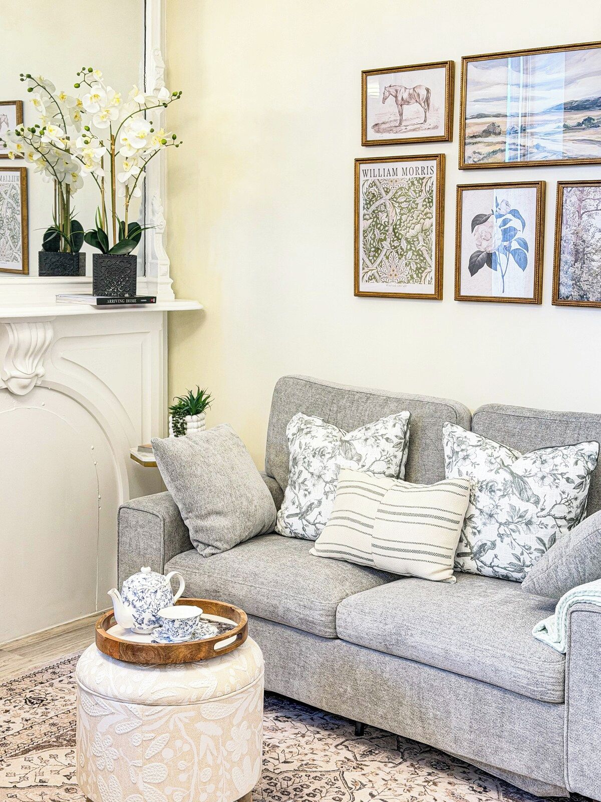 A living room with a grey sofa, floral cushions, a small round ottoman, a tray with teacups, and a fireplace with a white mantel.