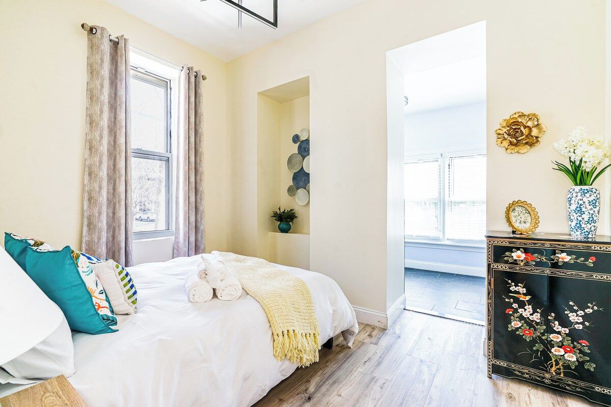 A bedroom with a bed, a dresser, a window with curtains, and a wall-mounted shelf with decorative items.