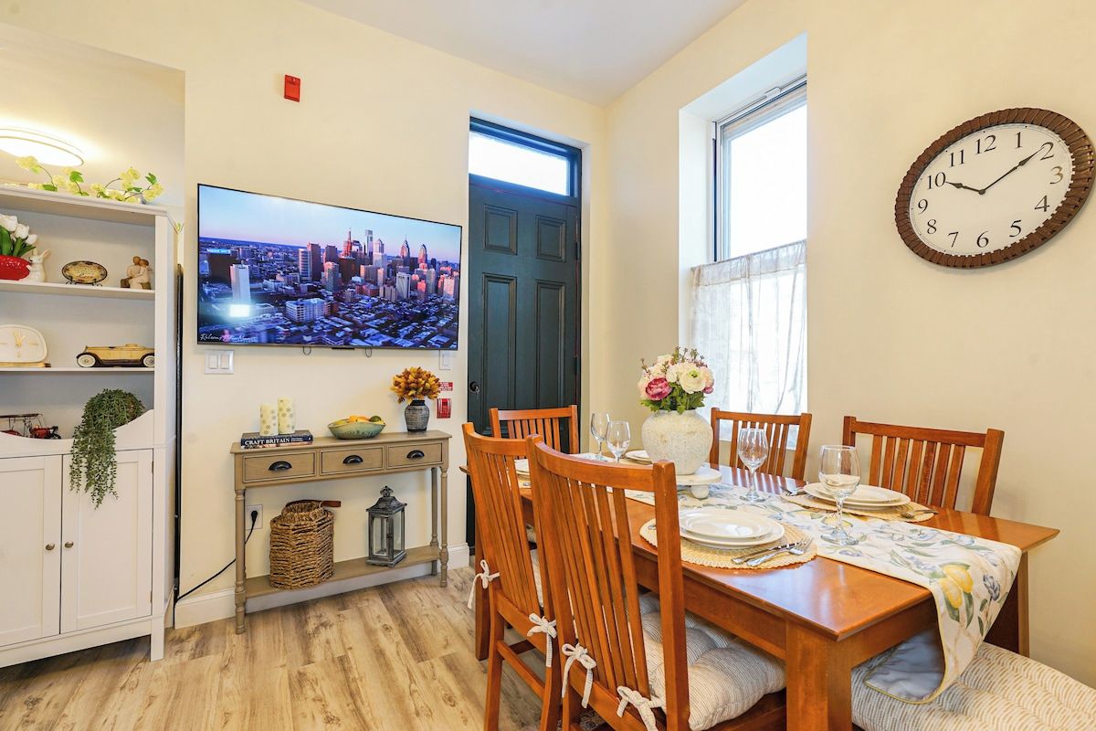 Un comedor con una mesa de madera preparada para cuatro personas, un televisor montado en la pared y un reloj en la pared.