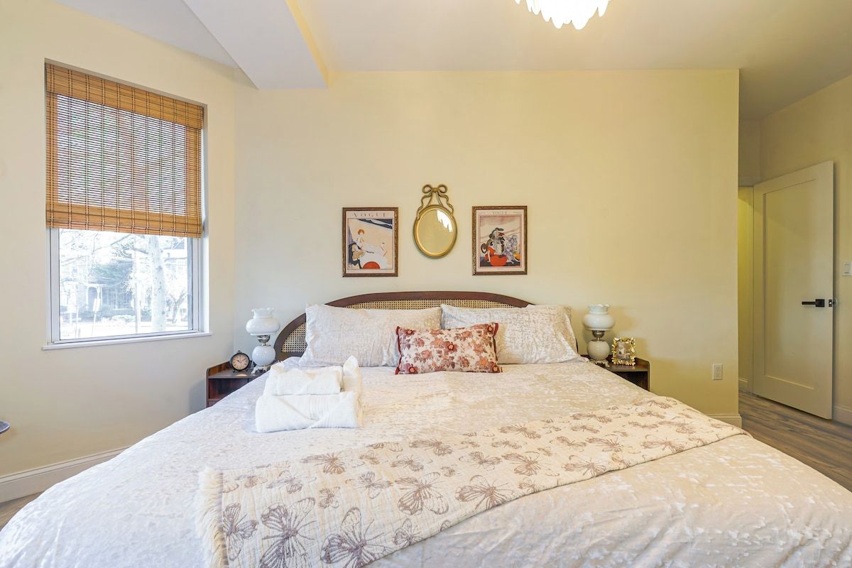 A bedroom with a large bed, a bedside table with a lamp, a window with blinds, and a door.