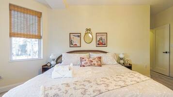 A bedroom with a large bed, a bedside table with a lamp, a window with blinds, and a door.