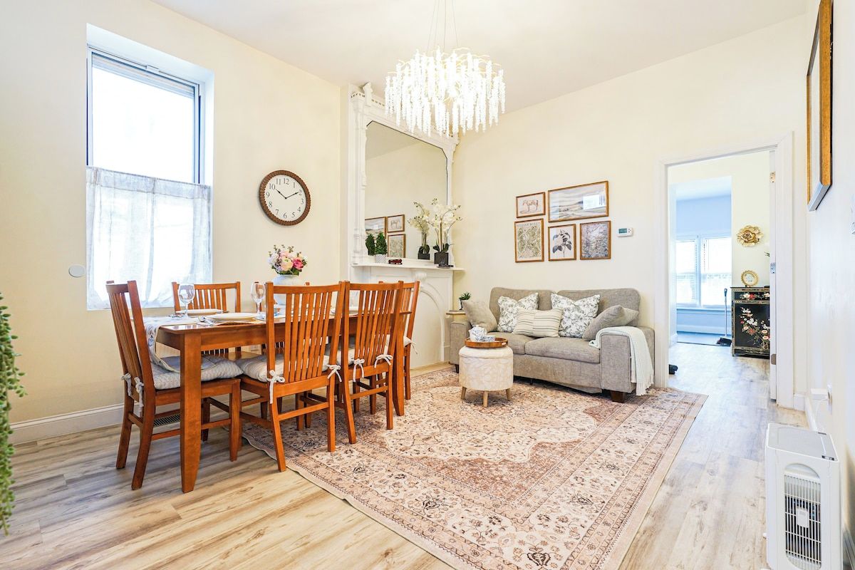 A dining area with a wooden table and chairs, a chandelier, a sofa, and a rug.