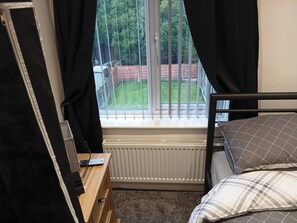 Et soveværelse med et vindue, en radiator, en trækommode, en seng med pude i tern og en sort gardin.