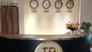 A reception desk with clocks labeled with city names, a logo, and a potted plant. - Tutto Passa Hotel (Tbilisi)