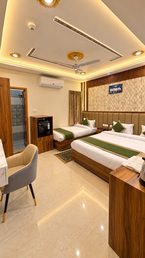 A hotel room with two beds, a desk, a chair, a television, and a ceiling fan. - BKRS Hotel (Varanasi)