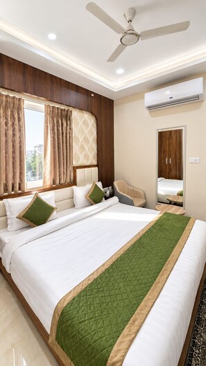 A hotel room with a large bed, a ceiling fan, an air conditioning unit, and a window with curtains. - BKRS Hotel (Varanasi)