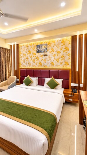 A hotel room with a large bed, a wooden nightstand, a desk, and a wall with a floral pattern. - BKRS Hotel (Varanasi)