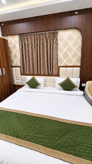 A hotel room with a large bed, a headboard with a patterned design, and a green quilted bedspread. - BKRS Hotel (Varanasi)