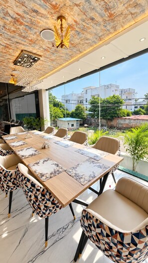 A modern dining area with a large table, patterned chairs, and a view of buildings through large windows. - BKRS Hotel (Varanasi)