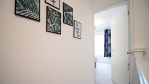 Ein Flur mit gerahmten Pflanzenbildern an der Wand, einer weißen Tür und Blick in einen Raum mit einem Fenster.