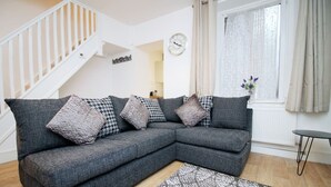 Ein Wohnzimmer mit grauem Ecksofa, gemusterten Kissen, einem Teppich mit Muster, einem kleinen Tisch, einer Treppe und einem Fenster mit Vorhängen.