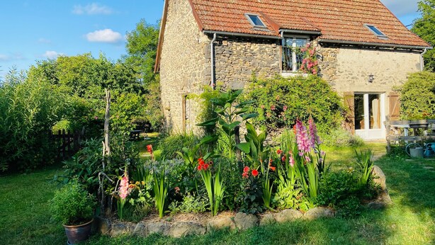 Ein Steinhaus mit Ziegeldach, ein Garten mit verschiedenen Pflanzen und Blumen sowie ein Holzzaun.