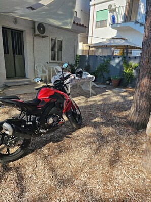 A red motorcycle parked in a gravel driveway, a white plastic table with chairs, and a potted plant. - Studio apartment with large garden. Fifty meters from the white beach of La Caletta. (La Caletta)