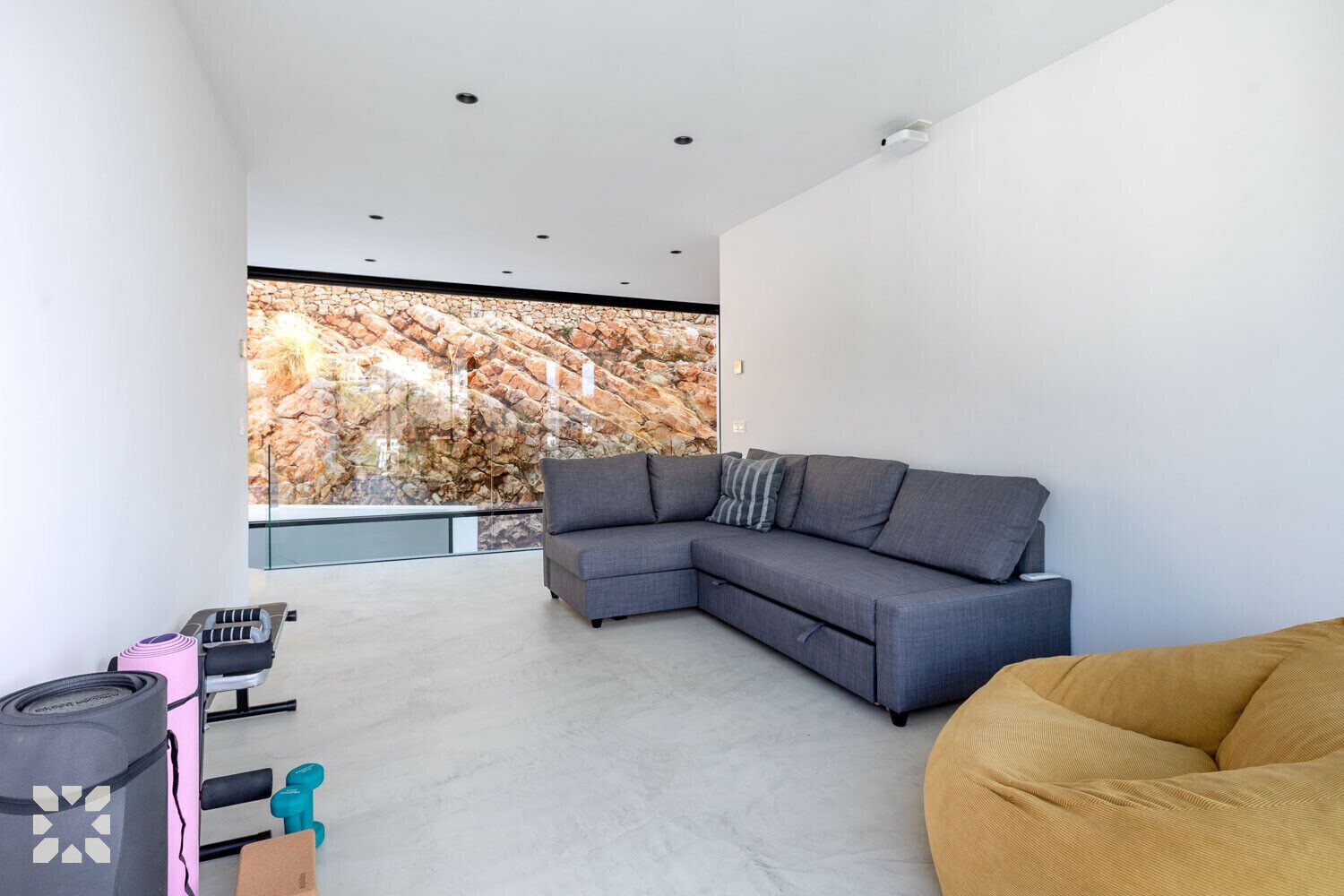 Un salon moderne avec un canapé d’angle gris, un fauteuil en forme de poire jaune et une grande fenêtre donnant sur un mur rocheux.