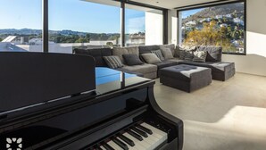 Een moderne woonkamer met een vleugelpiano, een hoekbank en grote ramen met uitzicht op een heuvelstad.