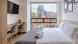 Ein modernes Hotelzimmer mit einem großen Bett, einem Schreibtisch, einem Sessel, einem Fernseher und Blick auf die Stadt.