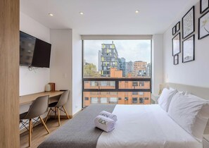 A modern hotel room with a large bed, a desk, a chair, a TV, and a view of the cityscape. - NOK Modern 1BR Near Parque 93 Bogota (Bogotá)
