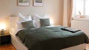 Un dormitorio con cama blanca, ropa de cama verde, dos cuadros enmarcados, una mesita de noche blanca y una ventana con cortinas.