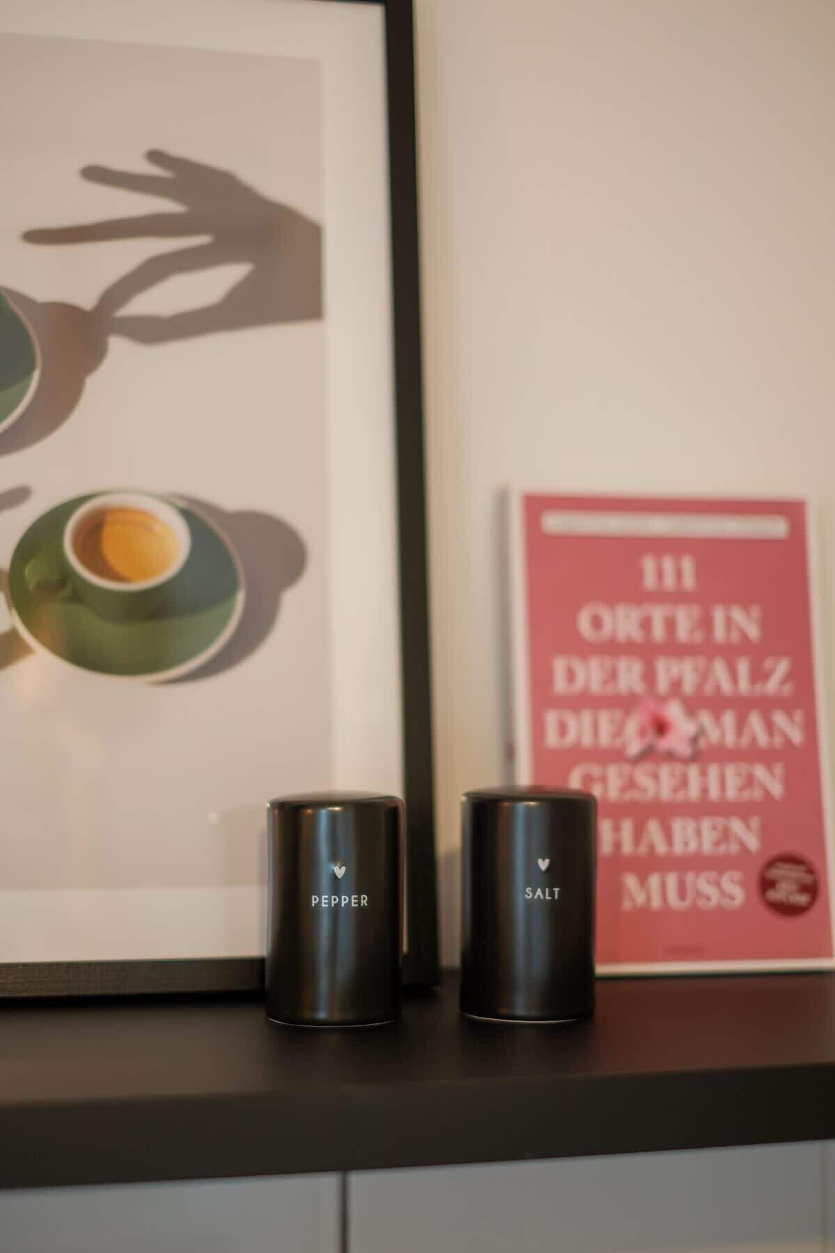 Een lijst met kunstwerk, twee zwarte recipiënten met de tekst 'PEPPER' en 'SALT', en een rode poster met tekst in een vreemde taal.
