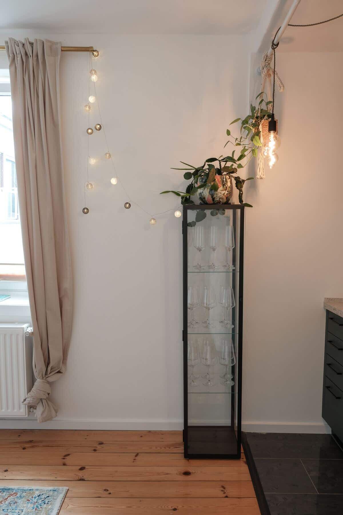 A room with a wooden floor, a glass cabinet with wine glasses, a plant with hanging lights, and a beige curtain.