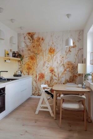 A modern kitchen with a white countertop, a wooden dining table, and a floral wallpaper. - Nestkoje - Ihr Ferienzuhause für die ganze Familie (Neustadt an der Weinstraße)
