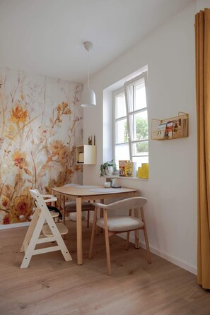 A dining area with a table and chairs, a window with curtains, and a wall with floral wallpaper. - Nestkoje - Ihr Ferienzuhause für die ganze Familie (Neustadt an der Weinstraße)