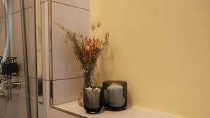 Ein Badezimmer mit einer Glasvase mit getrockneten Blumen auf einem Regal, einem Glaskerzenhalter und einem Metallhandtuchhalter.