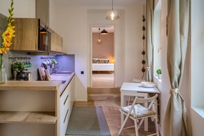 A modern kitchen with wooden cabinets, a dining area with a white table and chairs, and a bedroom with a bed and bedside lamp. - Exceptional Prague 1 Apt. in the City Center by Easybnb Premium (Prague)