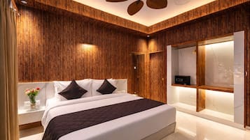 Une chambre à coucher moderne avec un mur recouvert de panneaux en bois, un lit avec une literie blanche et des oreillers foncés, une table de chevet ornée d’un vase de fleurs et un ventilateur de plafond.
