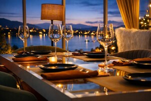 Mesa de comedor dispuesta con copas de vino, platos y una vela, con vistas a un cuerpo de agua y montañas.