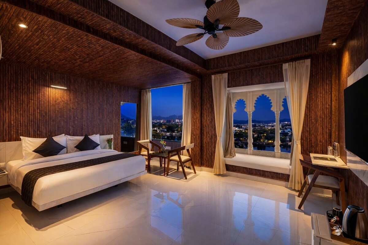 A modern hotel room with a large bed, a dining table, and a view of a cityscape.