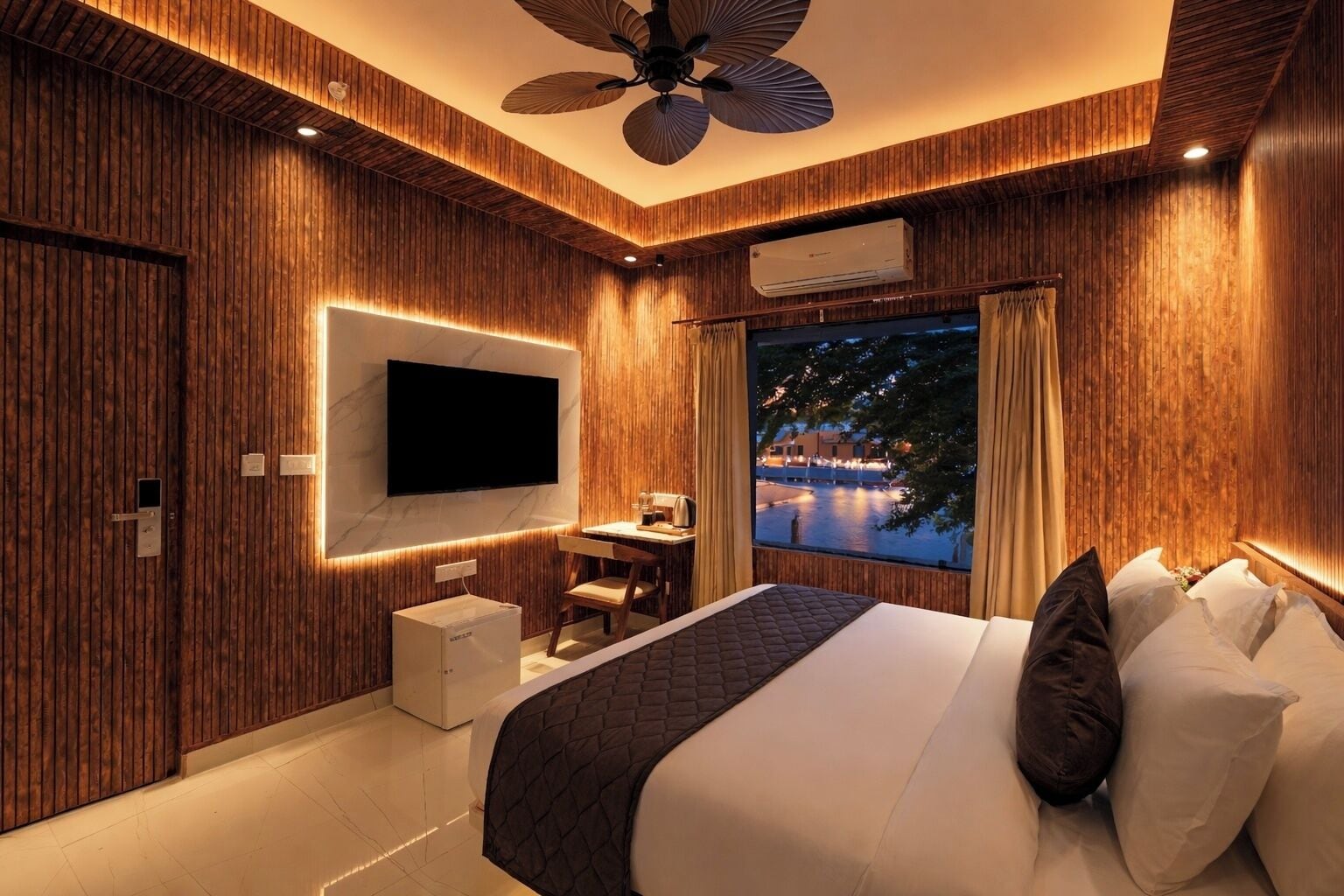 A modern bedroom with a large bed, a TV mounted on the wall, a desk with a chair, and a window with a view of a body of water.