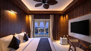 A hotel room with a large bed, a TV, a ceiling fan, and a view of a cityscape. - Shakuntalam Sagar - Luxury Boutique Lake View Hotel, Udaipur (Udaipur)