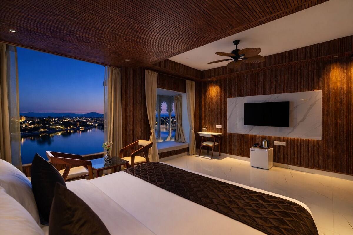 Una habitación de hotel moderna con una cama grande, un televisor montado en la pared, una zona de estar con mesa y sillas, y vistas a un paisaje urbano al atardecer.
