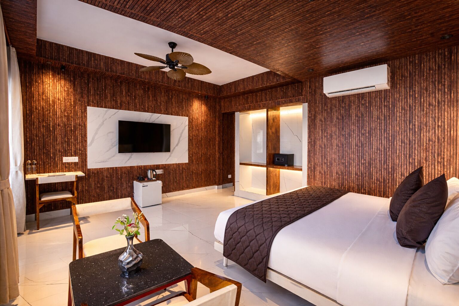 A modern hotel room with a bed, a desk with a chair, a TV mounted on the wall, and an air conditioning unit.
