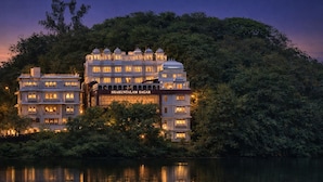 A multi-story building with a sign reading 'Shakuntalam Sagar' surrounded by dense greenery and reflecting on a calm body of water. - Shakuntalam Sagar - Luxury Boutique Lake View Hotel, Udaipur (Udaipur)