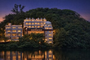 Un edificio de varios pisos con un letrero que dice 'Shakuntalam Sagar', rodeado de abundante vegetación y reflejado en una masa de agua tranquila.