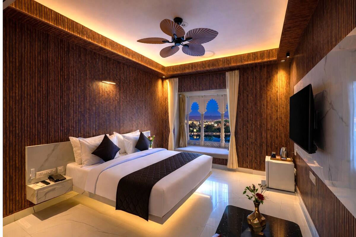 Habitación de hotel moderna con una cama grande, ventilador de techo, televisor y vistas a la ciudad al atardecer.