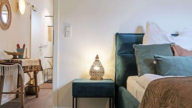 Ein Schlafzimmer mit Bett, Nachttisch, Lampe und Blick in einen weiteren Raum mit Tisch und Stühlen.