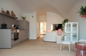 Una cocina moderna con electrodomésticos integrados, un comedor con mesa y silla blancos, y una sala de estar con sofá y televisor.