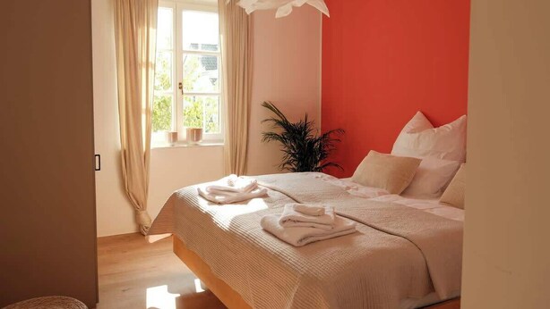 Un dormitorio con cama, cortinas en una ventana, una planta en maceta y una pared roja.