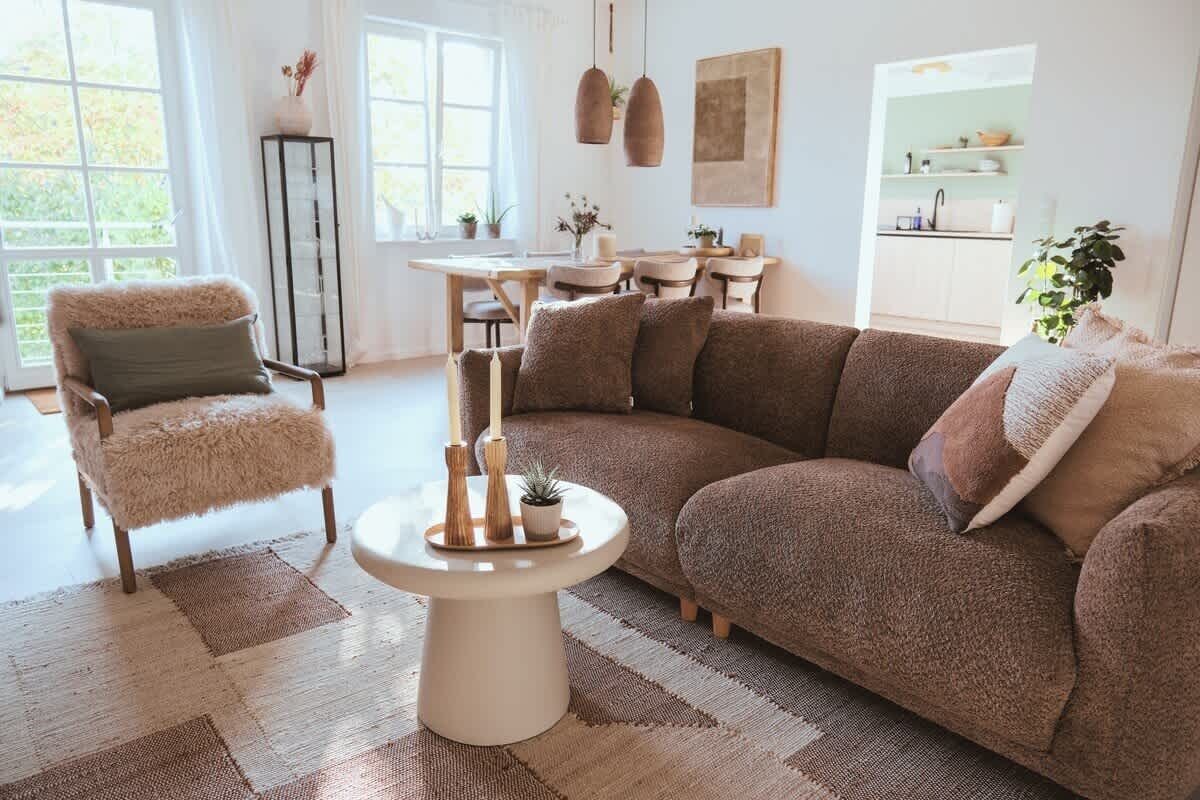 A cozy living room with a brown sofa, a fluffy chair, a round coffee table, and a view of a kitchen.
