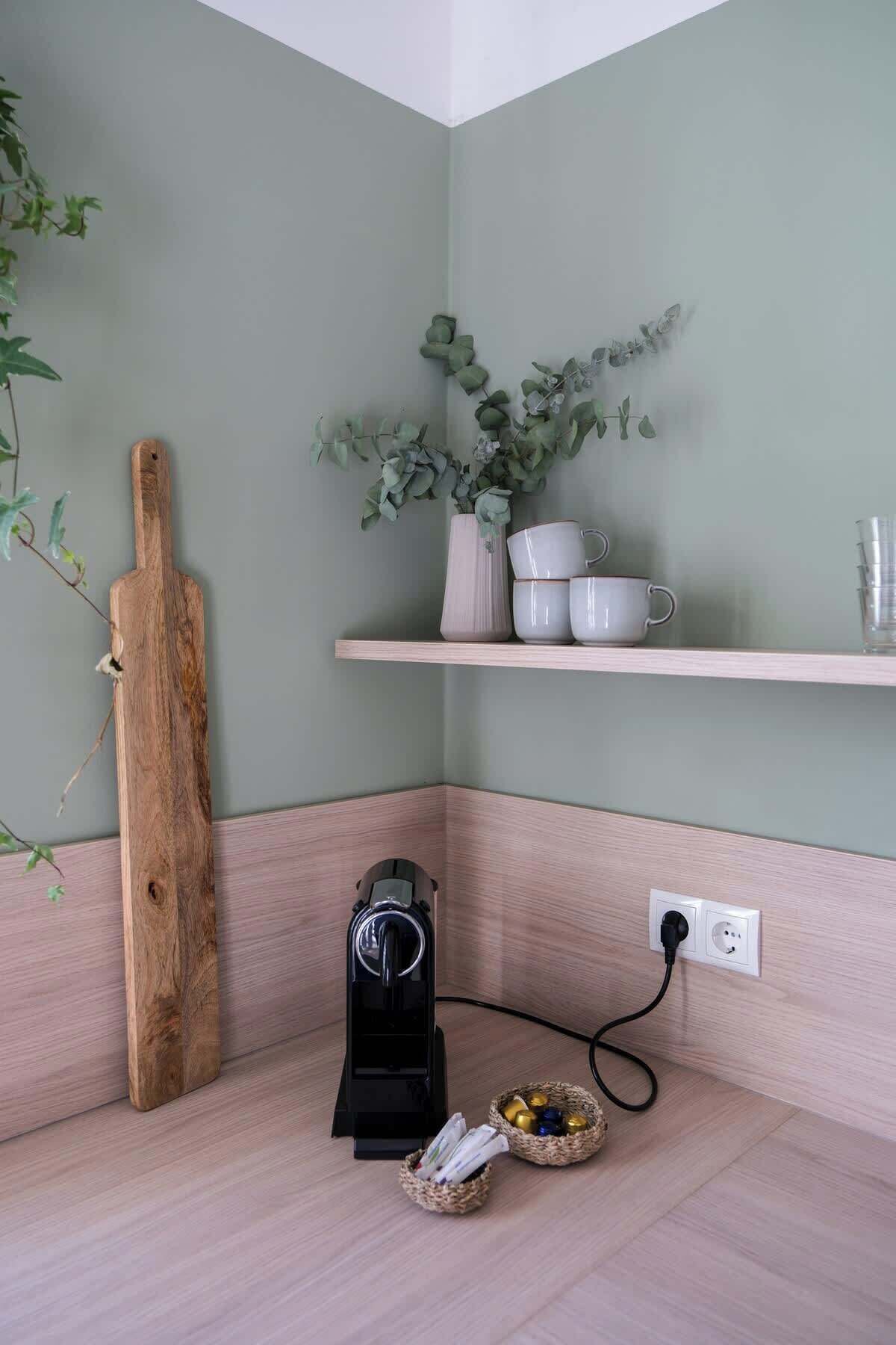 A corner of a room with a wooden cutting board, a coffee maker, a basket with snacks, and a shelf with cups and a vase with green leaves.