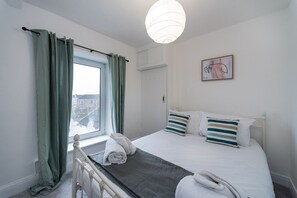 Ein Schlafzimmer mit einem Bett, einem Fenster mit Vorhängen, einem Nachttisch, einer Lampe und einem Bild an der Wand.