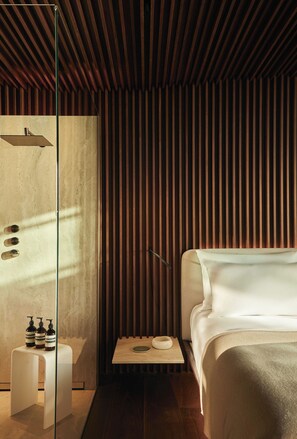 A modern hotel room with a wooden slatted wall, a glass shower enclosure, a bedside table with toiletries, and a bed with a white headboard. - Romeo Massa Lubrense (Massa Lubrense)