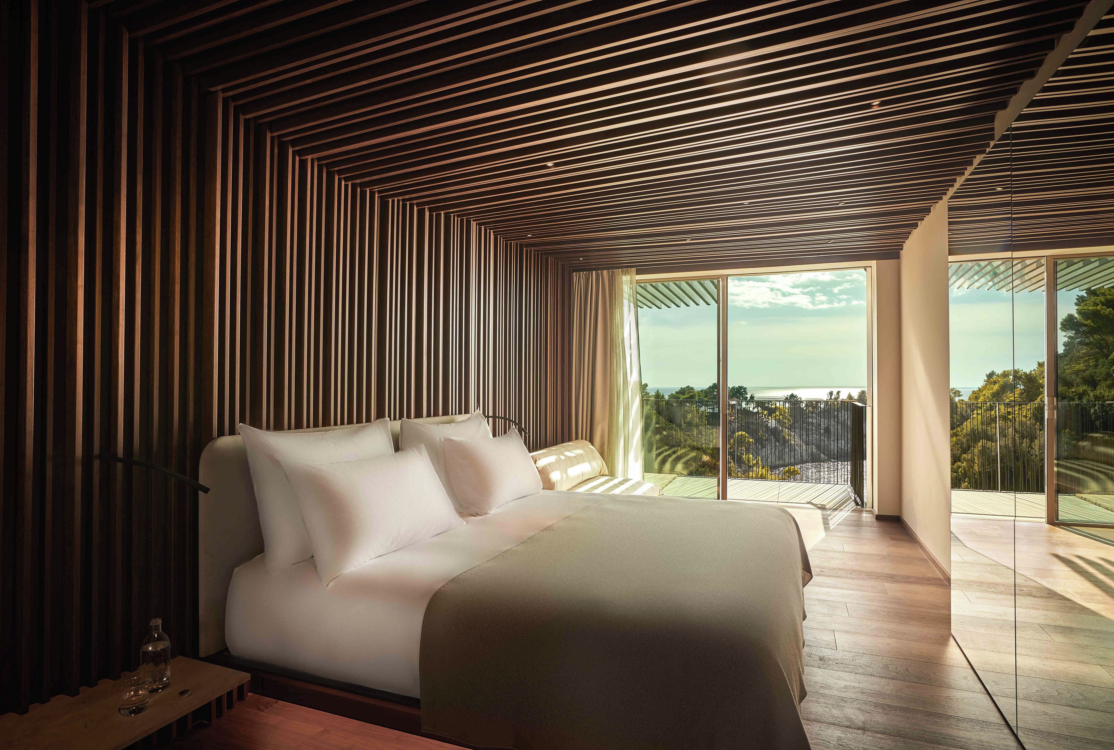 A modern bedroom with a large bed, a wooden slatted ceiling, and a balcony with a view of the sea.