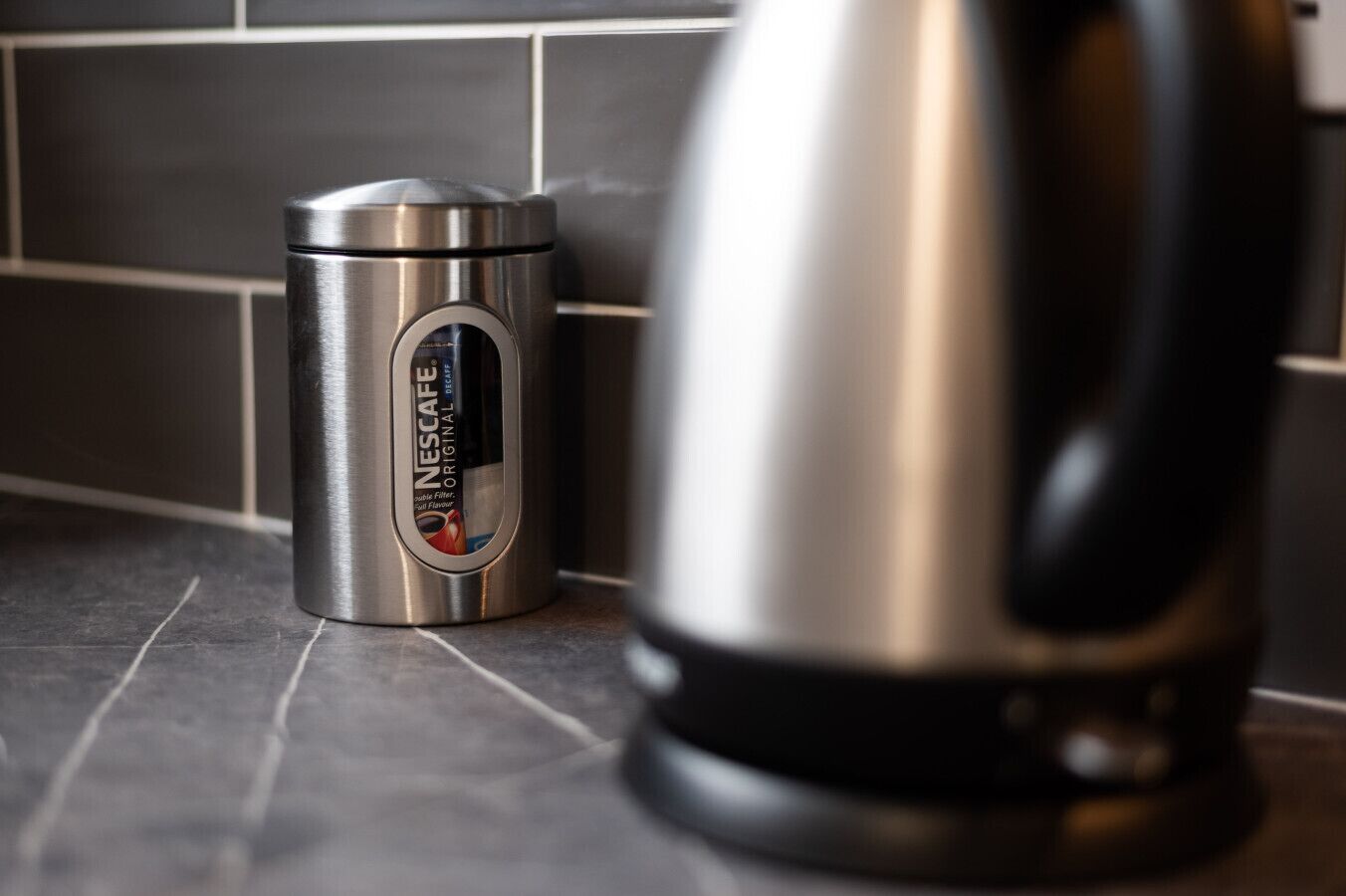 A stainless steel Nescafe coffee maker and a thermos on a tiled kitchen counter.