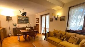 A living room with a sofa, a wooden coffee table, a dining table with chairs, a painting on the wall, and a lamp. - Casa Verocai Classic Retreat in Cortina (Cortina d'Ampezzo)