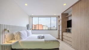 Ein modernes Hotelzimmer mit Bett, Nachttisch, Lampe, Fernseher und einem großen Fenster mit Blick auf die Stadt.